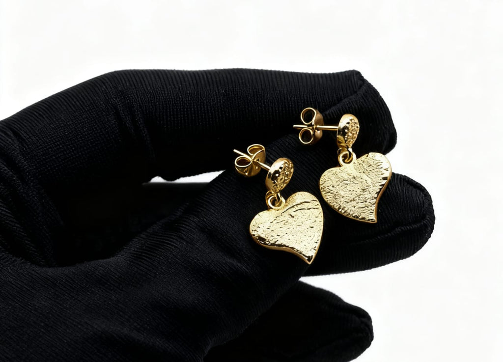 Gold heart-shaped earrings held by a black glove on a white background