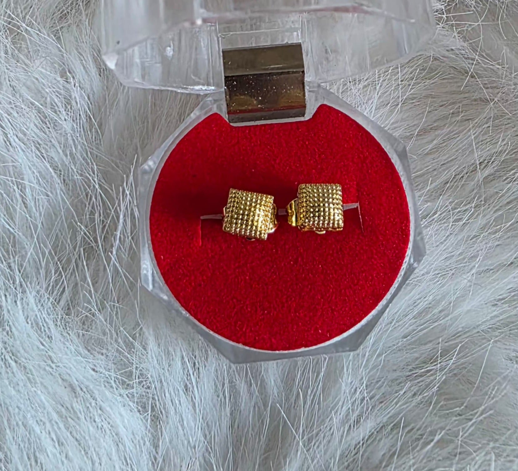 Gold square stud earrings in a clear jewelry box with red interior on a white fluffy surface.