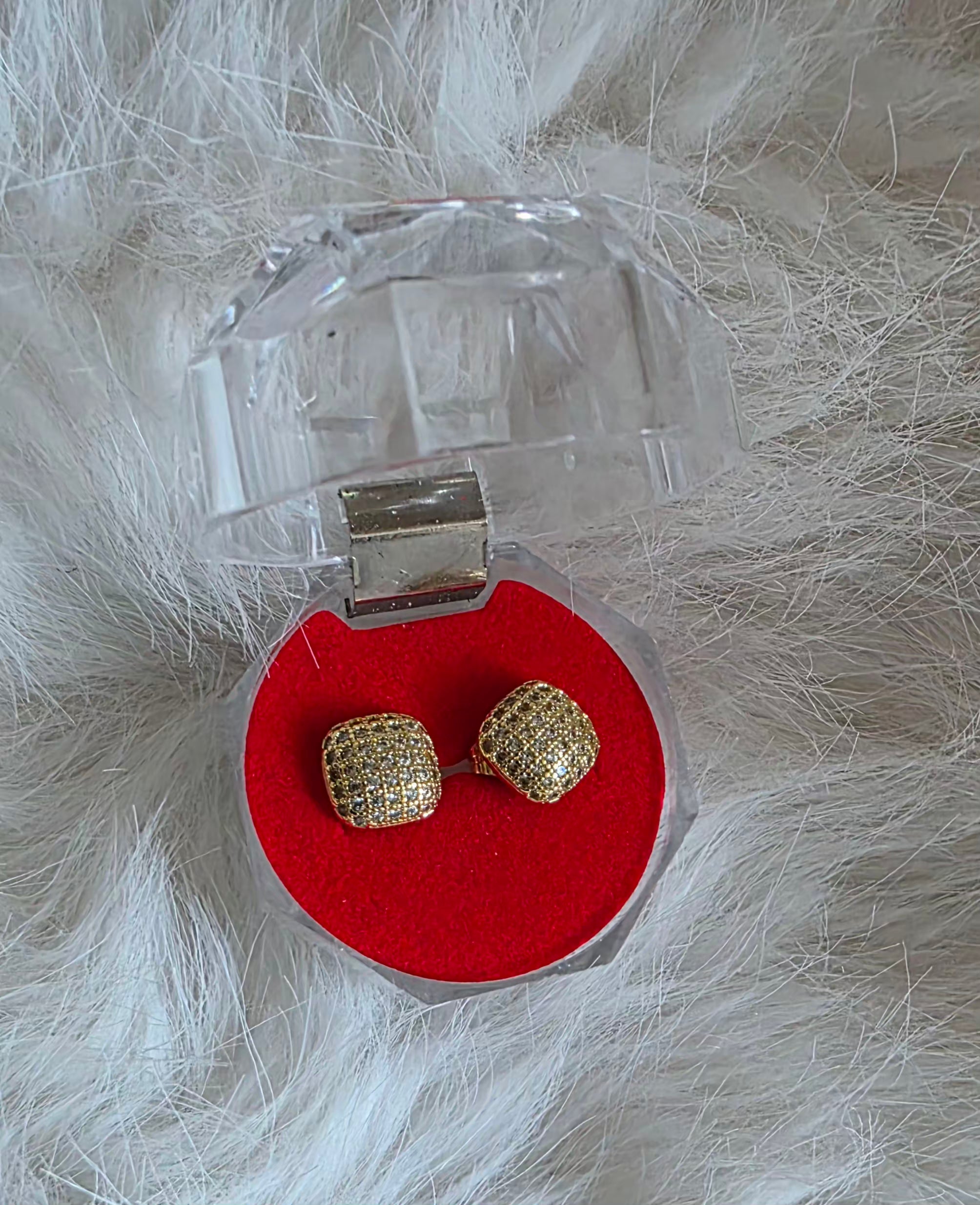 Gold earrings in a clear jewelry box on a white fluffy surface.